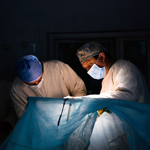 Ärzte im OP die einen Patienten unter einem gespannten blauen Abdecktuch behandeln. Sie tragen Mundschutz und Schutzkleidung sowie OP-Hauben